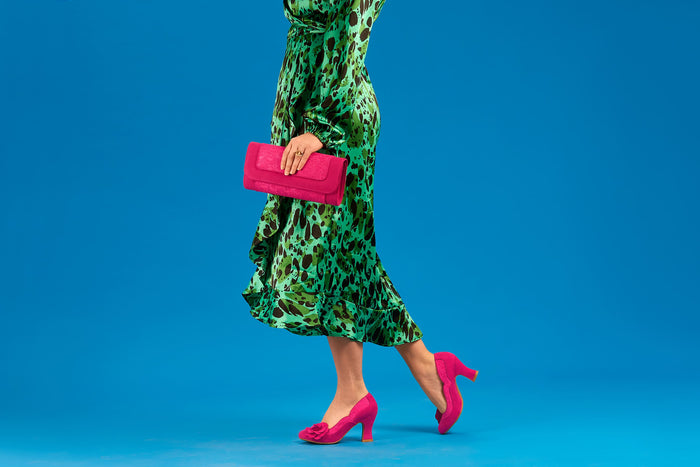 Person wearing a green patterned dress and pink shoes holding a pink clutch against a blue background
