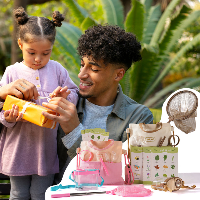 Two girls in meadow sitting on grass playing with ROBUD pink and green outdoor explorer kit accessories in warm golden afternoon light