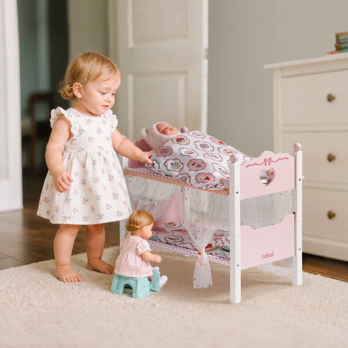 Little girl in a floral dress stands beside the ROBUD pink princess doll bunk bed placing a baby doll on the top bunk tier
