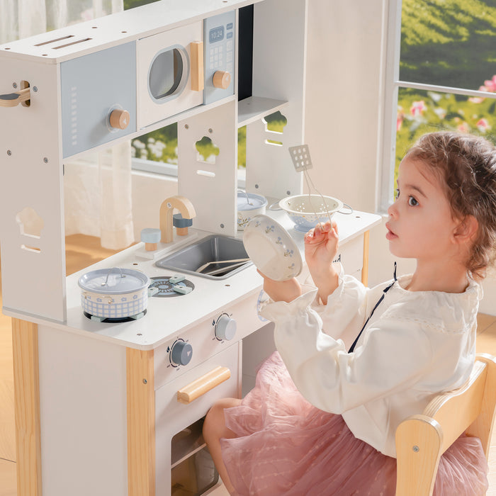 Girl seated beside the ROBUD white wooden play kitchen lifts a small cup while a pot rests on the stovetop burner.
