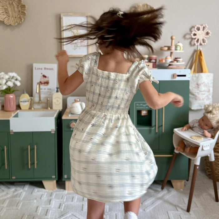 Girl in floral dress spinning with hair flying next to ROBUD green kitchen set in a warm cream and sage playroom