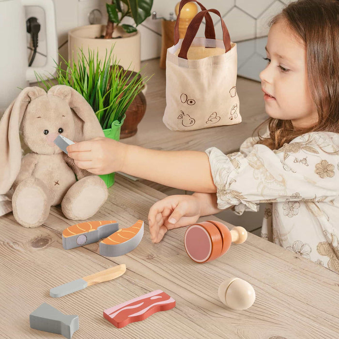 Young girl playing with ROBUD wooden meat cutting playset pieces including salmon, chicken leg, and fish beside a stuffed bunny toy at a wooden table
