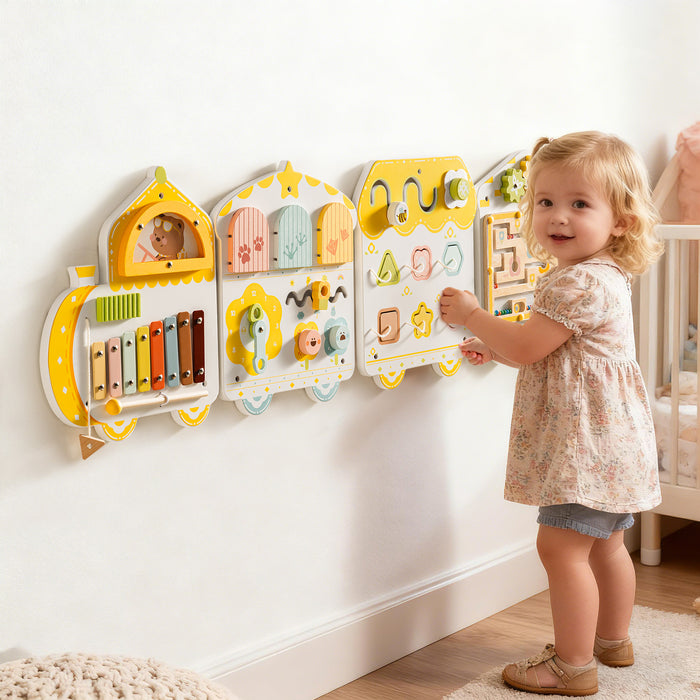 Toddler in a floral dress stands and plays at the ROBUD yellow spring train busy board mounted on a white nursery wall.