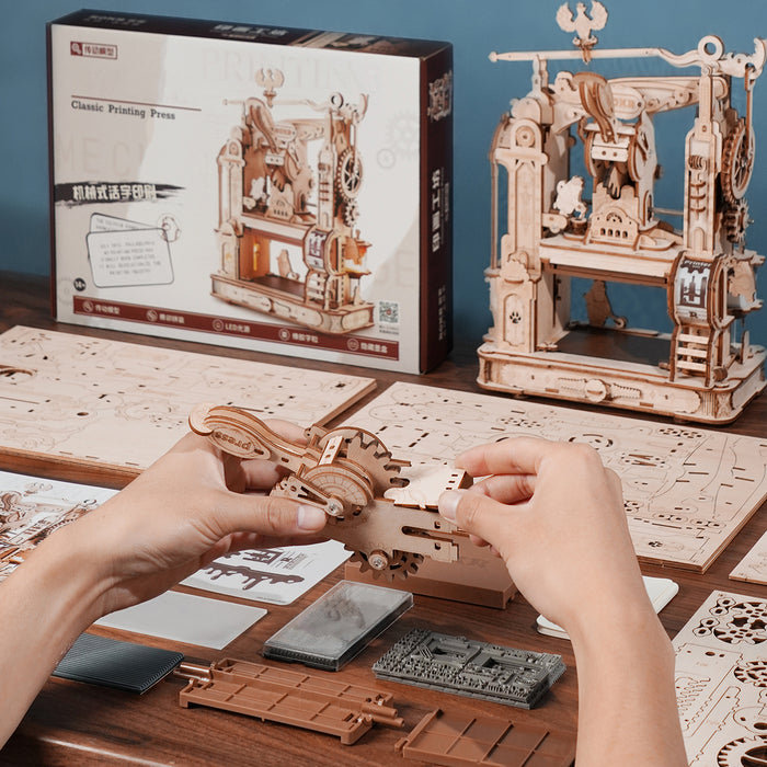 Hands assembling ROKR Classic Printing Press wooden puzzle on a desk with retail box and finished model in background