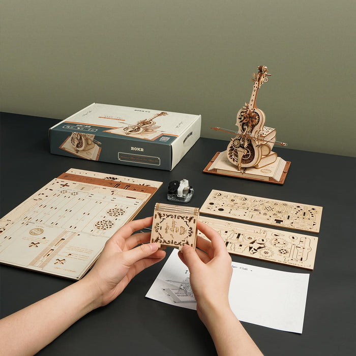Hands sorting laser-cut sheets beside assembled ROKR Magic Cello music box model and its retail packaging on dark table