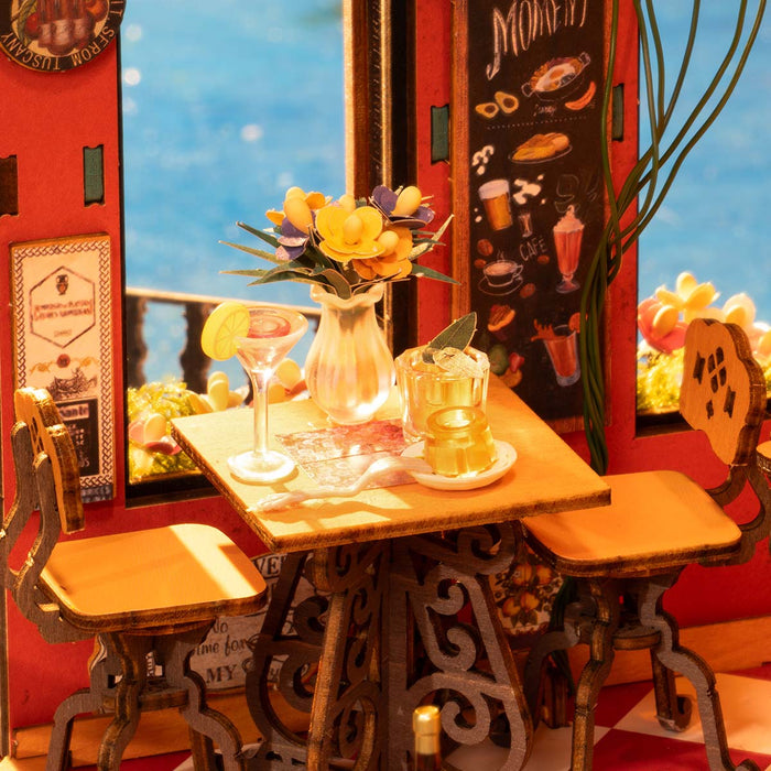 Rolife Cheers Bar DG173 miniature top-down interior view showing a cafe table with cocktail and flower vase, iron chairs, a chalkboard menu, and warm sunset lighting through the window