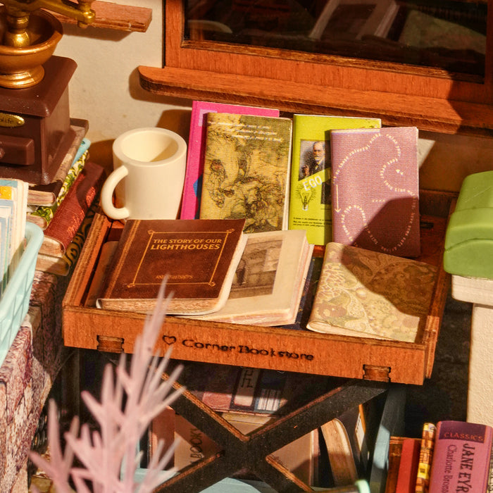Interior desk detail of the Rolife Corner Bookstore miniature with a tiny ceramic mug, stacked mini books, and a tray labeled Corner Bookstore