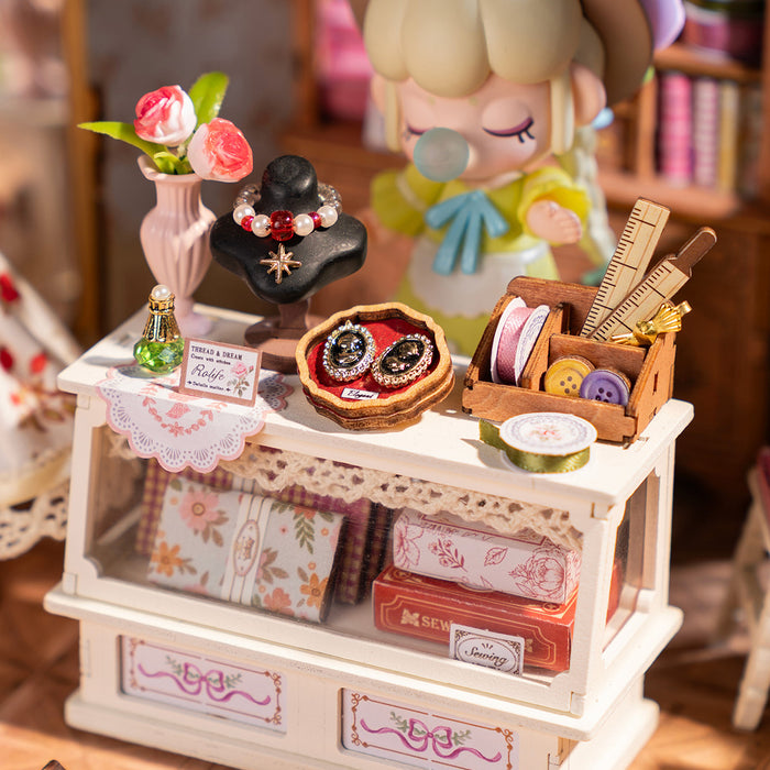 Rolife Elsa's Tailoring DG178 display counter close-up showing jewelry stand with necklace, brooch tray, buttons and tailor sewing box with Nanci figure watching