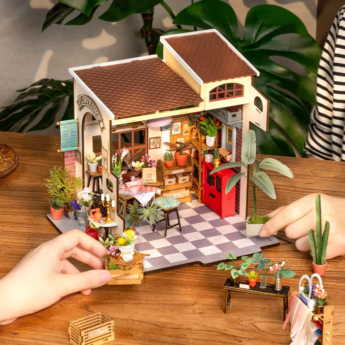 Rolife Emily's Flower Shop DG145 miniature house seen from the front on a wooden table as hands place small furniture pieces, with tropical plants beside it