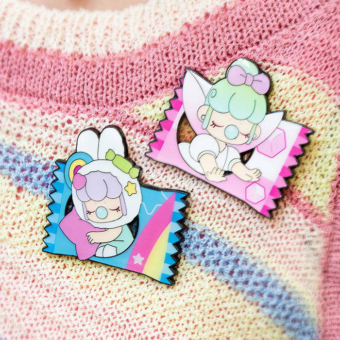 Two Rolife Nanci Sweet Time enamel pins on a rainbow-striped knit sweater displaying the Fluffy Cloud blue candy wrapper and the Tea Cup pink candy wrapper designs