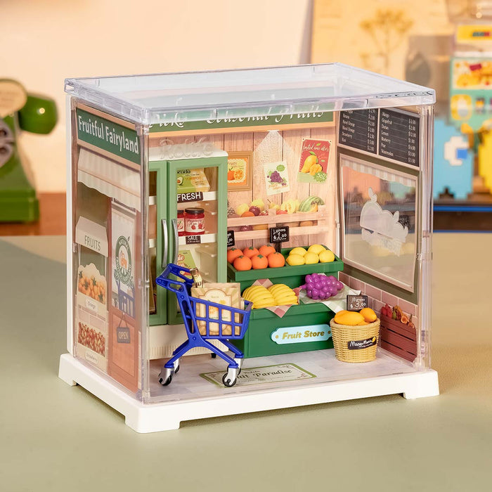 Rolife Nature Energy Fruits DWS01B lifestyle view showing the green fruit store miniature with pushable shopping cart inside its clear acrylic display case on a desk.