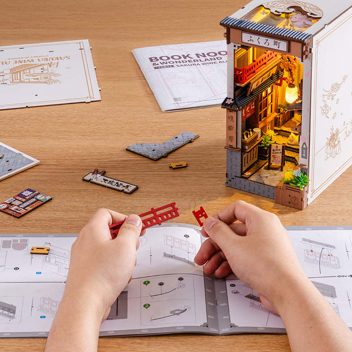 Rolife DIY miniature book nook TGB11 Sakura Wine Alley assembly scene with hands following printed instructions beside the illuminated Japanese alleyway diorama