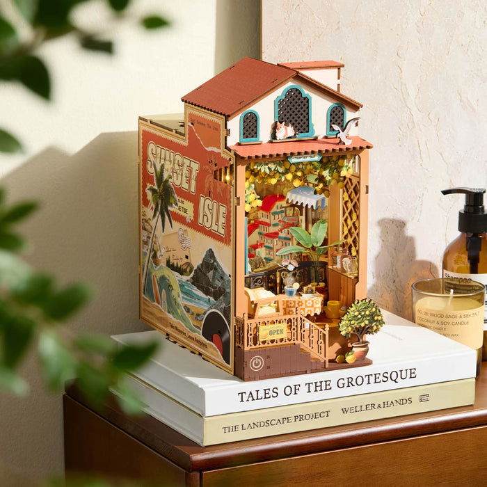 Rolife assembled book nook model placed on a wooden desk next to a book, showing a Mediterranean house facade with a lemon tree garden and neon sign