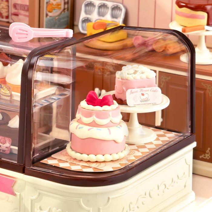 Rolife Tasty Cake Shop DIY miniature house DW019B displayed at an angle showing the fully assembled pink bakery interior with glass display counter, cakes, and warm LED lighting.