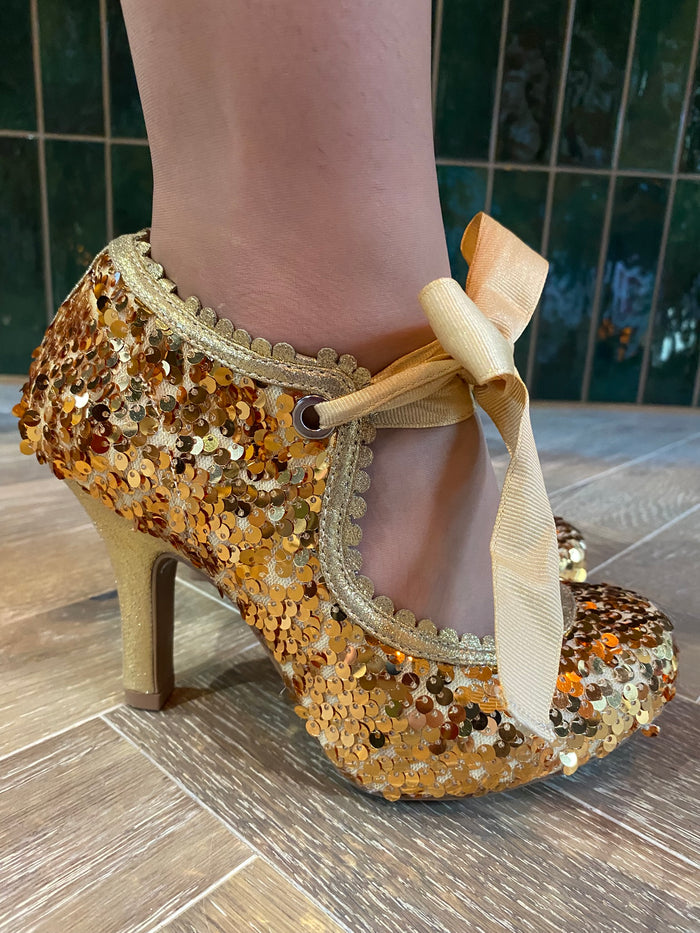 Gold sequin high-heeled shoe with a bow on a tiled floor.